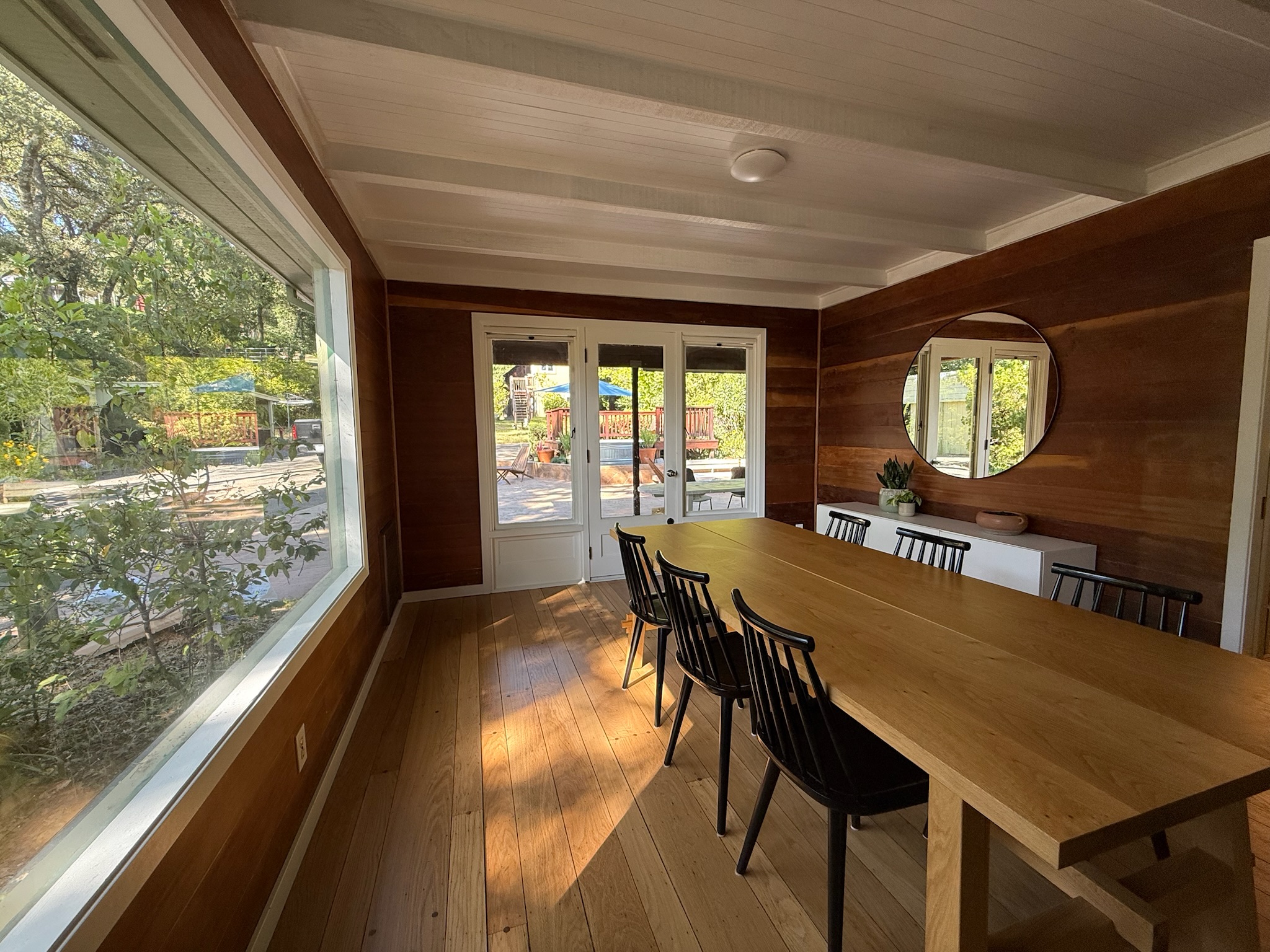 A sun-drenched view of the dining room, where a single door flanked by tall windows leads to the expansive backyard patio. Light dances through the canopy of mature trees outside.