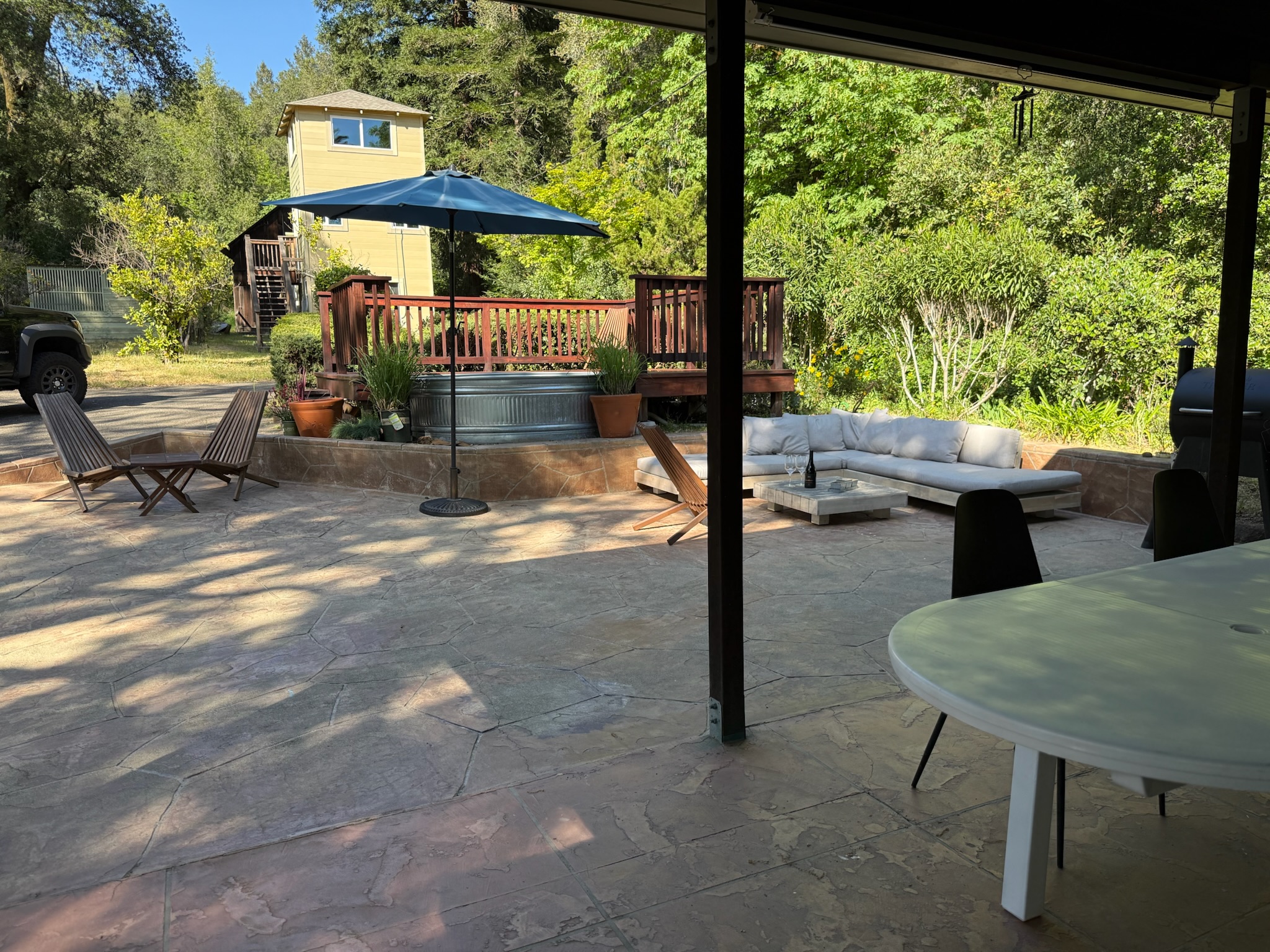The spacious patio is built for gathering and relaxing, featuring a shaded outdoor lounge, a Traeger grill, and plenty of open space framed by lush greenery.